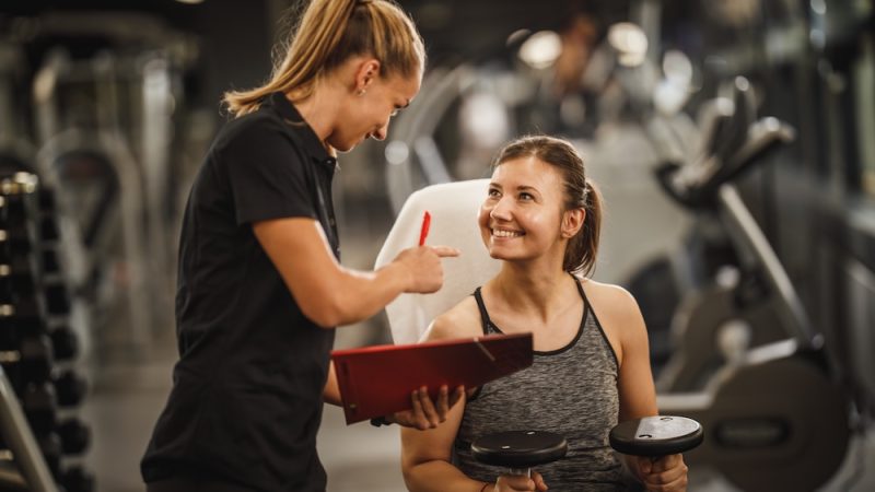 Shot of a muscular young woman in sportswear working out with female personal trainer at the gym. She is pumping up her muscule with dumbbell.