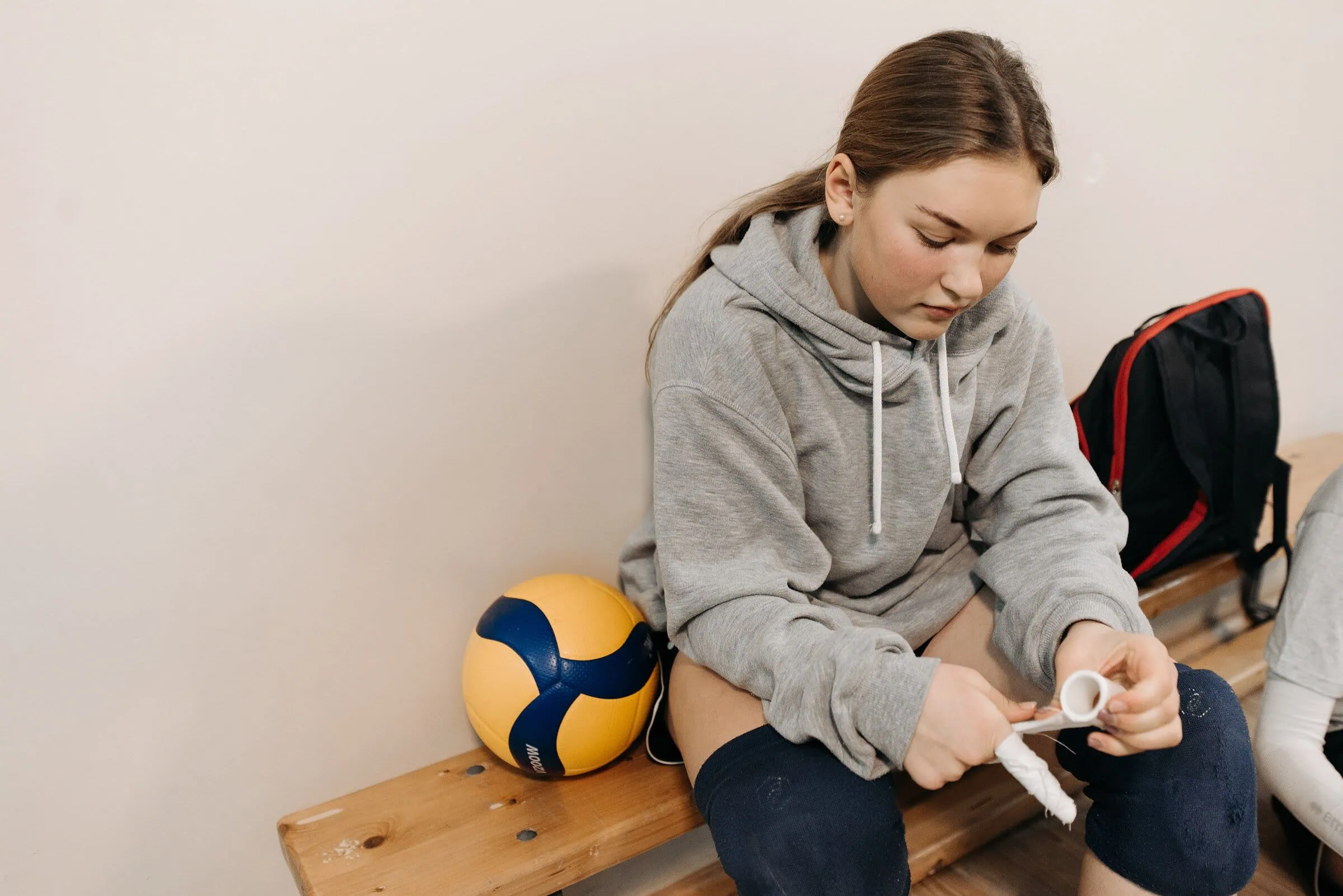 Young female athlete in gray hoodie wrapping hands with tape, sitting on bench with volleyball.
