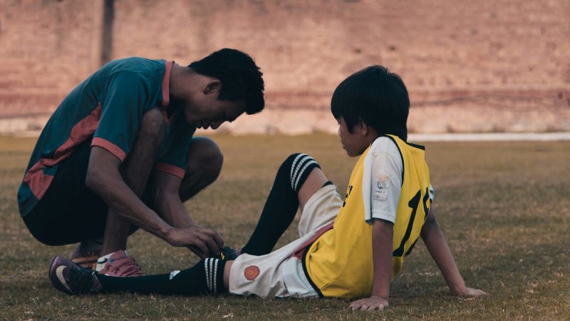 Coach kneeling to check the ankle of an injured youth soccer player sitting on the field
